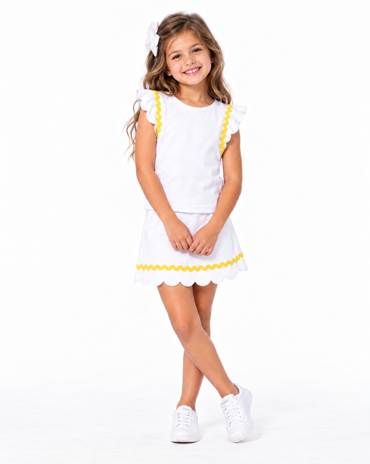 A young girl with long light brown hair wearing a white two-piece outfit with yellow scalloped trim, featuring a short-sleeve top with ruffle details and a matching skirt. She is also wearing a white hair bow and white sneakers, standing against a solid w