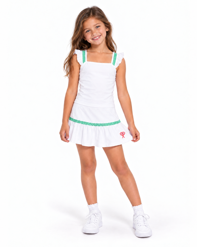 A young girl modeling a white sleeveless tennis top and skirt with green patterned shoulder straps and a green patterned waistband, paired with white socks and white sneakers, on a solid white background.