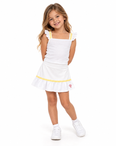 A young girl with long light brown hair stands against a white studio background wearing a white sleeveless top and skirt with yellow trim and white sneakers.