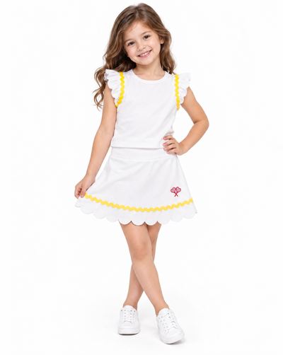 A young girl with long brown hair stands against a white studio background wearing a white sleeveless dress with yellow ruffle trim, a yellow scalloped hem, and a small red logo, paired with white sneakers.