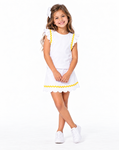 A young girl with long light brown hair wearing a white two-piece outfit with yellow scalloped trim, featuring a short-sleeve top with ruffle details and a matching skirt. She is also wearing a white hair bow and white sneakers, standing against a solid w